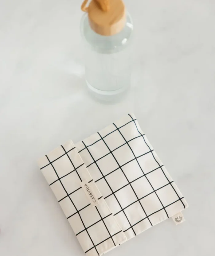 L'Enveloppe à Encas de Casavida, pochette multiusage en coton à carreaux à côté d'une gourde en verre